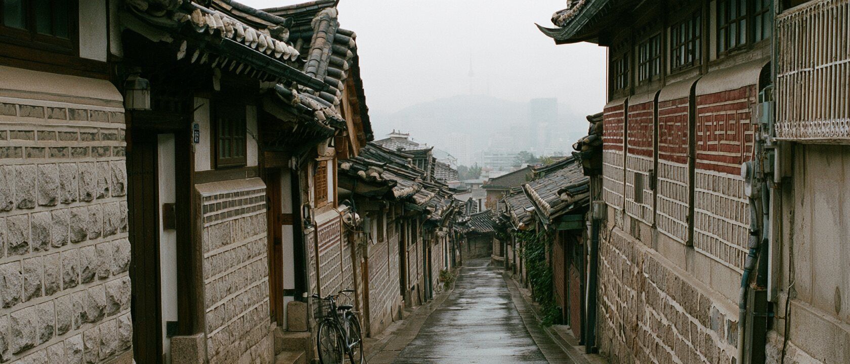 A quiet Bukchon alley in Seoul on an overcast morning: stone walls, hanok roofs stepping down the hill, a parked bicycle, Namsan tower faint in the haze